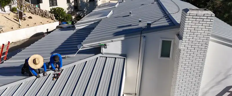 Two roofers seaming light gray standing seam panels beside a white brick chimney