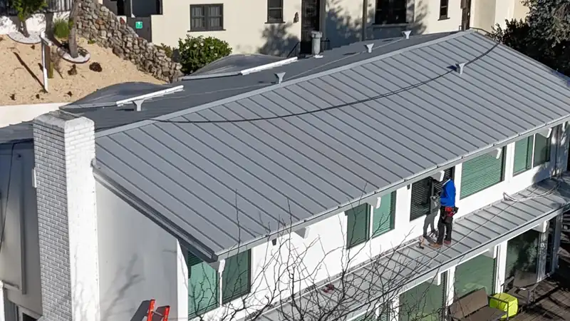 Roofer fitting standing seam panels at the lower eave of a white house with green shutters