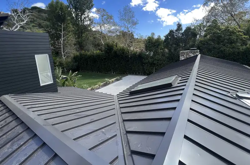 Black standing seam metal roof planes meeting at a hip, with a skylight and a stone chimney