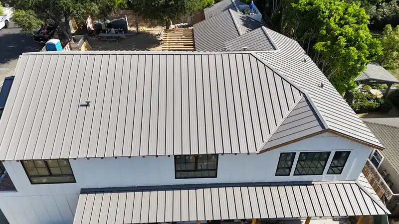 Aerial view of a gray standing seam metal roof with hips and a gable over a white board-and-batten house